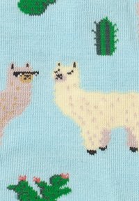 Zwei Lamas, eins beige und eins cremefarben, mit grünen Kakteen und Pflanzen auf einem hellblauen, gestrickten Stoffhintergrund.