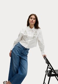 Camicia bianca corta con ricami floreali argento decorativi sul davanti, abbinata a jeans a gamba larga blu a vita alta. Sedia nera pieghevole nelle vicinanze.