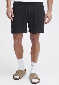 Schwarze Baumwoll-Shorts mit strukturierter Oberfläche, ausgestattet mit zwei Fronttaschen und einem verstellbaren Kordelzugbund. Kombiniert mit weißen Socken und Sandalen.
