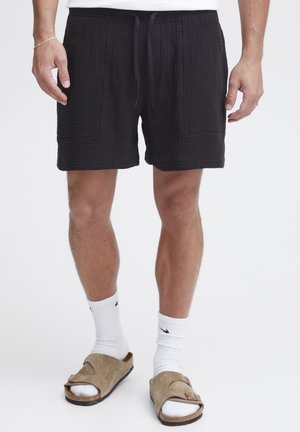 Black cotton shorts with a textured surface, featuring two front pockets and an adjustable drawstring waistband. Worn with white socks and sandals.