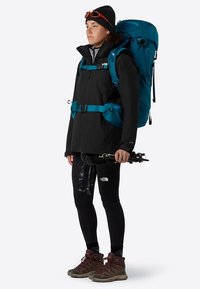 Czarna wodoodporna kurtka z turkusowymi akcentami, czarne legginsy, brązowe buty trekkingowe i duży turkusowy plecak. Trzyma kijki trekkingowe.