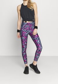 Svart ärmlös topp med en knut vid midjan, kombinerad med färgglada leggings i ett rosa och blått leopardi-mönster, och svarta sneakers.