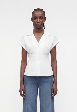 Frau mit geflochtenem Haar trägt eine figurbetonte weiße Kurzarmbluse mit Knöpfen und weite blaue Jeans, steht vor einem schlichten Hintergrund.