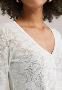 Gilet en tricot blanc doté d'un délicat motif en dentelle, d'un col en V et de cinq boutons à l'avant. Texture douce avec un design léger.