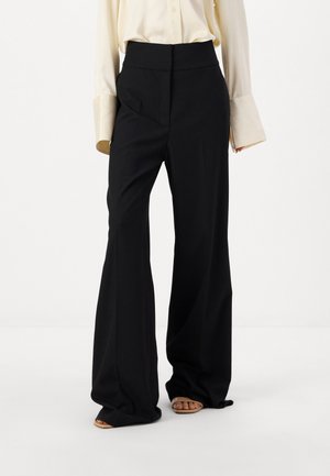 Pantalons larges noirs à taille haute, fabriqués en tissu lisse. Présentent un pli avant et des poches, associés à une chemise à manches longues de couleur claire.