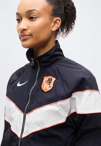 Νεαρή γυναίκα που φοράει αθλητικό μπουφάν με φερμουάρ σε μαύρο και άσπρο, με πορτοκαλί έμβλημα CFCL και λευκό λογότυπο Nike.