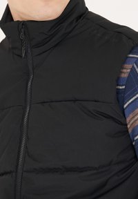 Schwarze Daunenweste mit hohem Kragen, ausgestattet mit einem Reißverschluss und einer gesteppte Textur, kombiniert mit einem blau-braunen karierten Hemdärmeln.