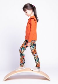 Pomarańczowa koszula z długim rękawem z karbowanym dekoltem, połączona z czarnymi legginsami w kwiaty z pomarańczowymi, zielonymi i białymi wzorami. Lekkie białe buty sportowe.