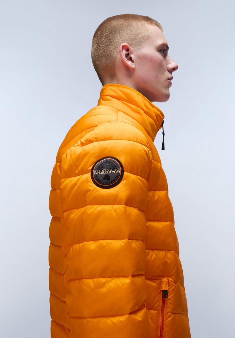 Pomarańczowa kurtka puchowa z wysokim kołnierzem, pikowany wzór, kieszeń na zamek i okrągła czarna naszywka z logo na lewym ramieniu.