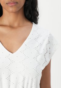 Bílý top s výstřihem do V, opatřený výšivkou z perliček, květinovým vzorem a krátkými rukávky. Měkká textura s kulatými výřezy a detaily.