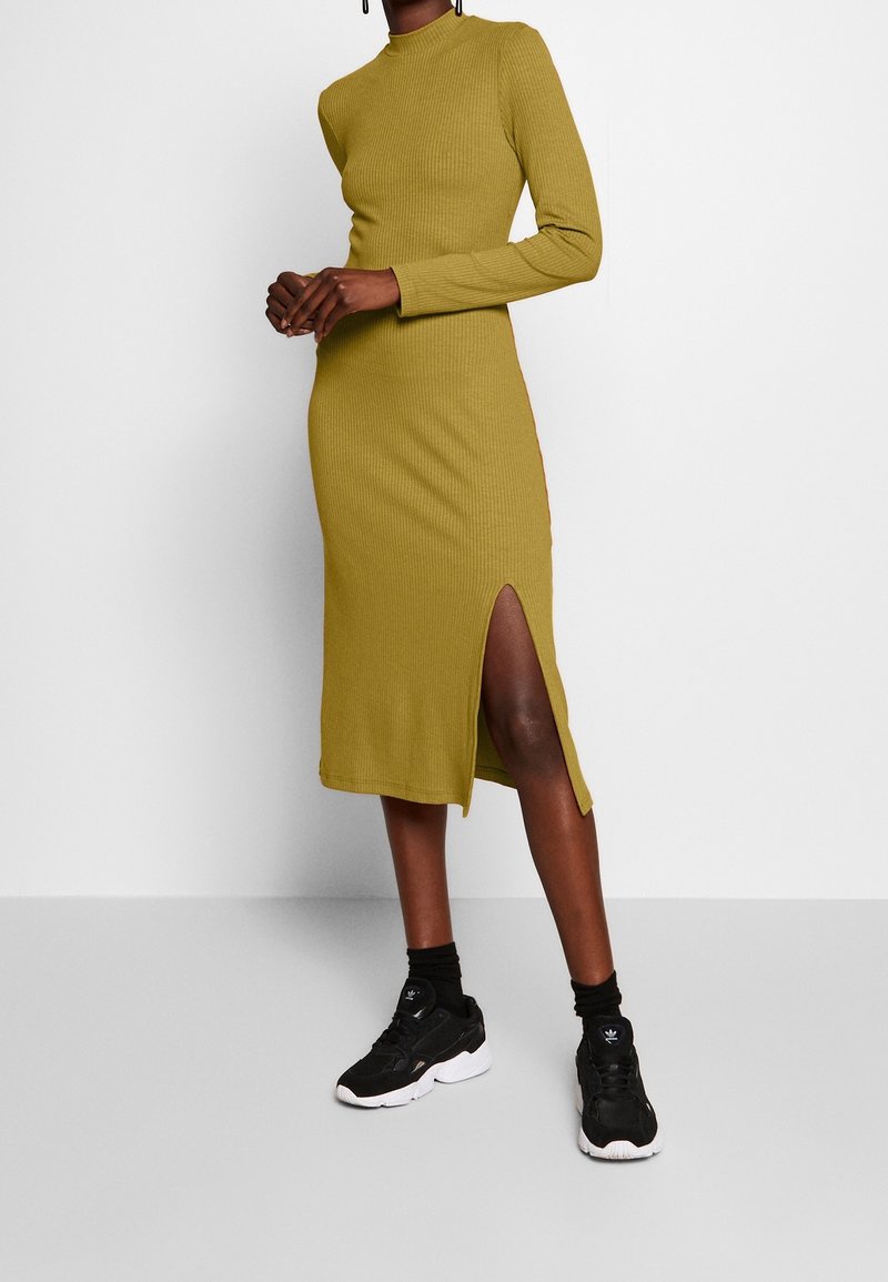 Robe côtelée vert moutarde avec un col cheminée et des manches longues, dotée d'une fente latérale haute jusqu'à la cuisse. Associée à des chaussures de sport noires.