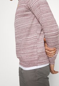 Maglione a righe rosse chiare con maniche lunghe. Il materiale sembra morbido e testurizzato. Orlo bianco visibile nella parte inferiore. Nessun logo o design visibile.