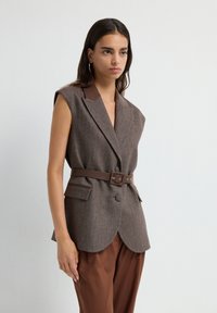 Brun herringbone ærmeløs blazer med brun krave, figursyet taljebælte og frontlommer, båret over brune bukser.
