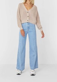 Licht beige geribbelde gebreide cardigan met diepe V-hals en drie grote knopen, gecombineerd met lichtblauwe high-waisted wijde jeans en sneakers.