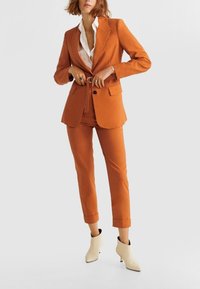 Blazer orange et pantalon fuselé assorti, avec une chemise blanche en dessous. Comprend une ceinture, deux poches avant et des bottines crème.
