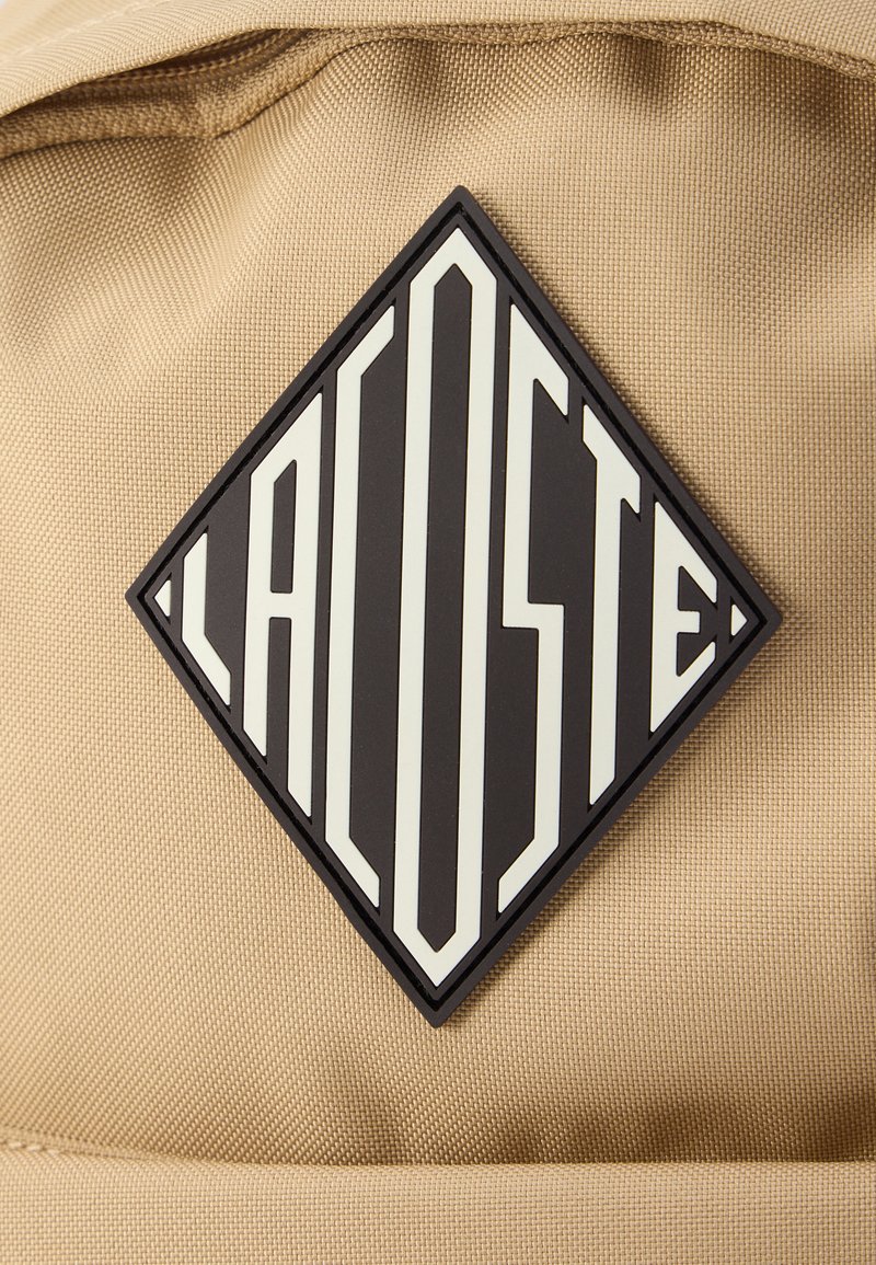 Tela beige con un parche de goma negro en forma de diamante que presenta "LACOSTE" en letras blancas verticales y en negrita, junto con acentos geométricos.