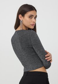 Szary, prążkowany top z długim rękawem o skróconym fasonie. Materiał ma delikatny połysk. Modelka ma długie kolczyki i jest lekko obrócona w bok.