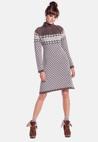 Kobieta ubrana w brązowo-białą dzianinową sukienkę w wzory z długimi rękawami i golfem, zestawioną z brązowymi botkami za kostkę z podwiniętymi mankietami.