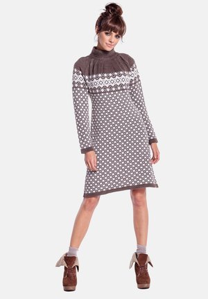 Kobieta ubrana w brązowo-białą dzianinową sukienkę w wzory z długimi rękawami i golfem, zestawioną z brązowymi botkami za kostkę z podwiniętymi mankietami.