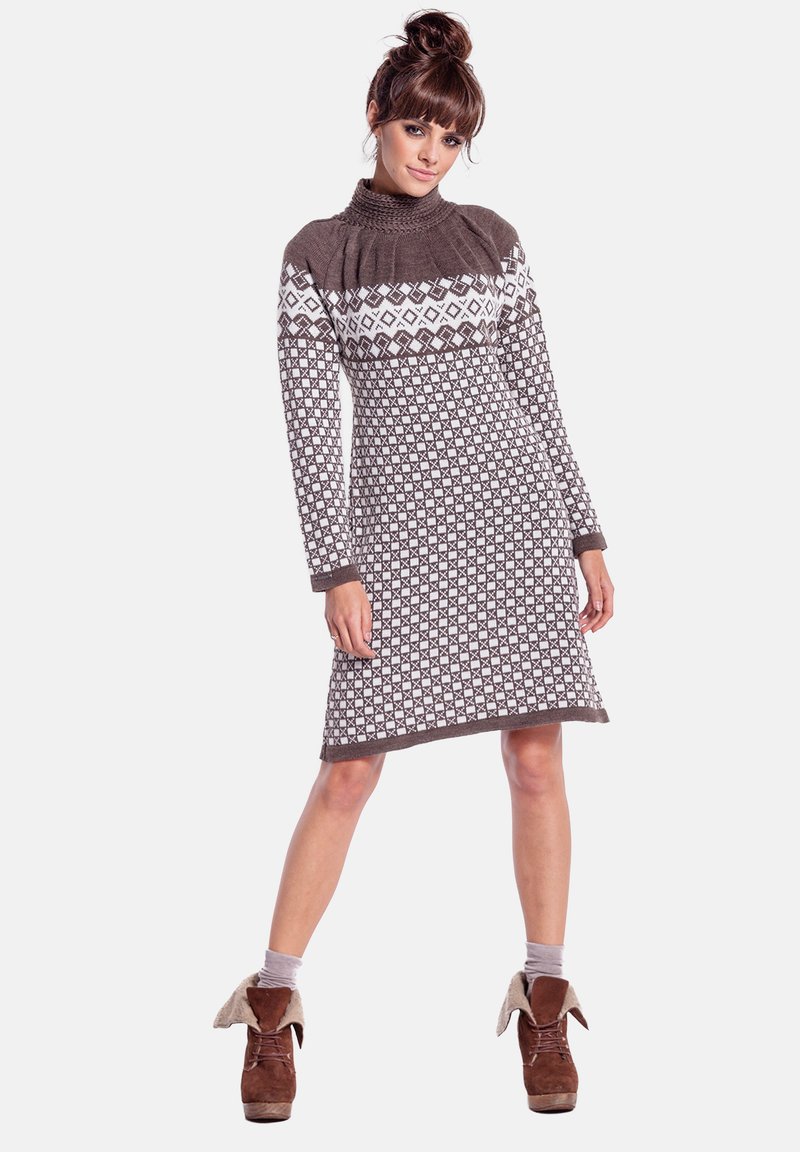 Kobieta ubrana w brązowo-białą dzianinową sukienkę w wzory z długimi rękawami i golfem, zestawioną z brązowymi botkami za kostkę z podwiniętymi mankietami.