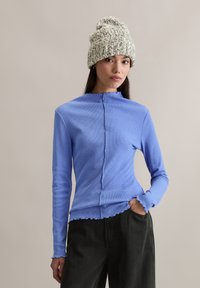 Blaues, geripptes Langarmshirt mit hohem Kragen, zentraler Naht und gezackten Kanten. Kombiniert mit schwarzen, locker sitzenden Hosen.