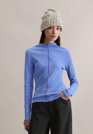 Niebieska prążkowana bluzka z długim rękawem i wysokim kołnierzem, z centralnym szwem i karbowanymi krawędziami. W zestawie z czarnymi, luźnymi spodniami.