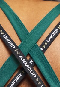 Zielone ramiączka sportowego biustonosza krzyżują się na tle w kolorze skóry, z czarnymi elastycznymi paskami z napisem „UNDER ARMOUR” w białym kolorze i złotymi akcentami.
