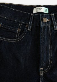 Jean taille haute bleu foncé avec coutures visibles, poche avant, bouton en métal et étiquette indiquant la taille et les détails d'origine.