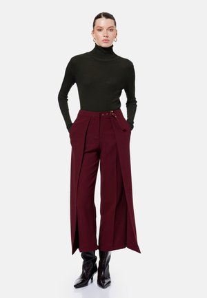 Frau trägt schwarzen gerippten Rollkragenpullover, bordeauxrote weite Hosen mit Gürtel und vorderen Schlitzen, sowie schwarze spitze Stiefeletten, steht mit Händen in den Taschen.