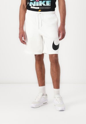 Weiße Nike-Shorts mit schwarzem Logo am linken Bein, aus weichem Stoff gefertigt; kombiniert mit weißen Sportschuhen und weißen Kurzsocken.