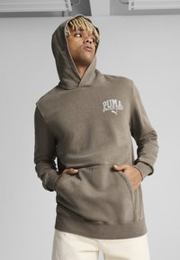 Sudadera marrón con bolsillo canguro, textura suave y un logo blanco que dice "PUMA sports club". Presenta un corte relajado y capucha.