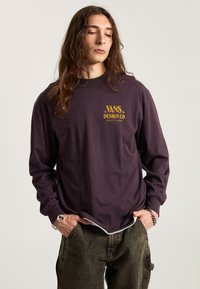 Fioletowa koszula z długim rękawem z napisem "VANS DESIGN CO." w złocie z przodu, zestawiona z luźnymi, teksturowanymi ciemnymi spodniami.