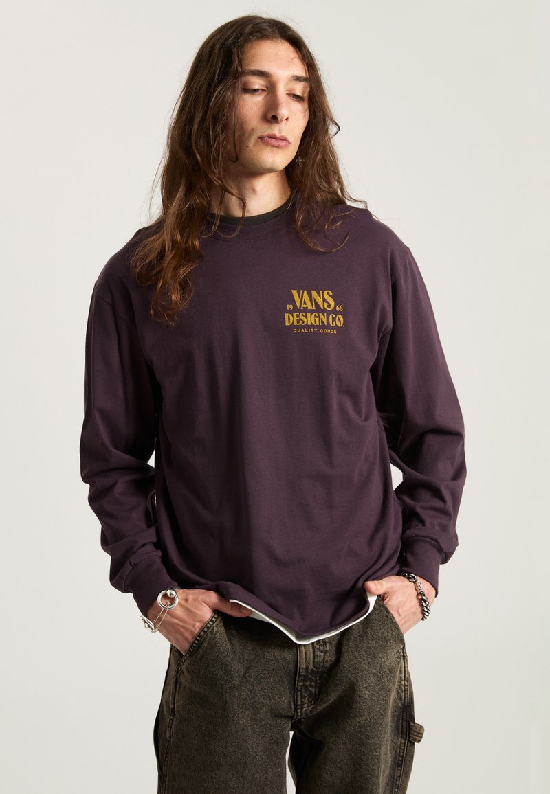 Fioletowa koszula z długim rękawem z napisem "VANS DESIGN CO." w złocie z przodu, zestawiona z luźnymi, teksturowanymi ciemnymi spodniami.