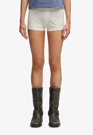 Persoon die een korte beige broek met kleine bloemenprint draagt, een blauw gestreept overhemd en donkergrijze leren laarzen tot halverwege de kuit, rechtop staat.