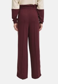 Pantaloni larghi di colore marrone bordeaux realizzati in tessuto morbido, con vita elasticizzata e dettagli con pieghe frontali. Vista posteriore che mette in risalto la semplicità del design.