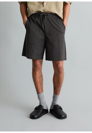 Man draagt zwarte short met trekkoord, staat met handen in de zakken, gecombineerd met lichtgrijze sokken en zwarte instappers.