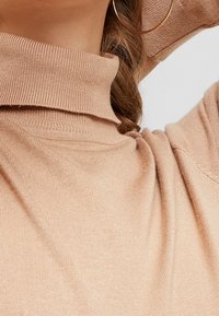 Femme portant un pull à col roulé beige et une grande boucle d'oreille en or, avec les cheveux partiellement visibles sur un fond neutre.