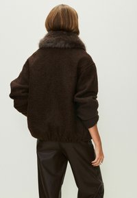 Chaqueta de piel sintética marrón con una textura suave, hombros caídos y dobladillo acanalado. El cuello está forrado con un pelaje más oscuro. Combinada con pantalones de cuero marrón.