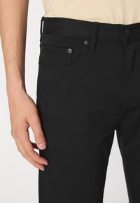 Jean en denim noir à coupe slim, design à cinq poches, avec des coutures contrastantes. Comprend des accessoires en métal et une texture lisse.