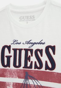 T-shirt in cotone biologico bianco con grafica rossa e blu navy. Il design frontale presenta la scritta "Guess" e "Los Angeles" in testo ricamato con un accento grafico.