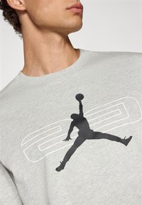 Camiseta de algodón gris claro con una gran silueta negra de un jugador de baloncesto en pleno salto, con acentos gráficos blancos detrás.