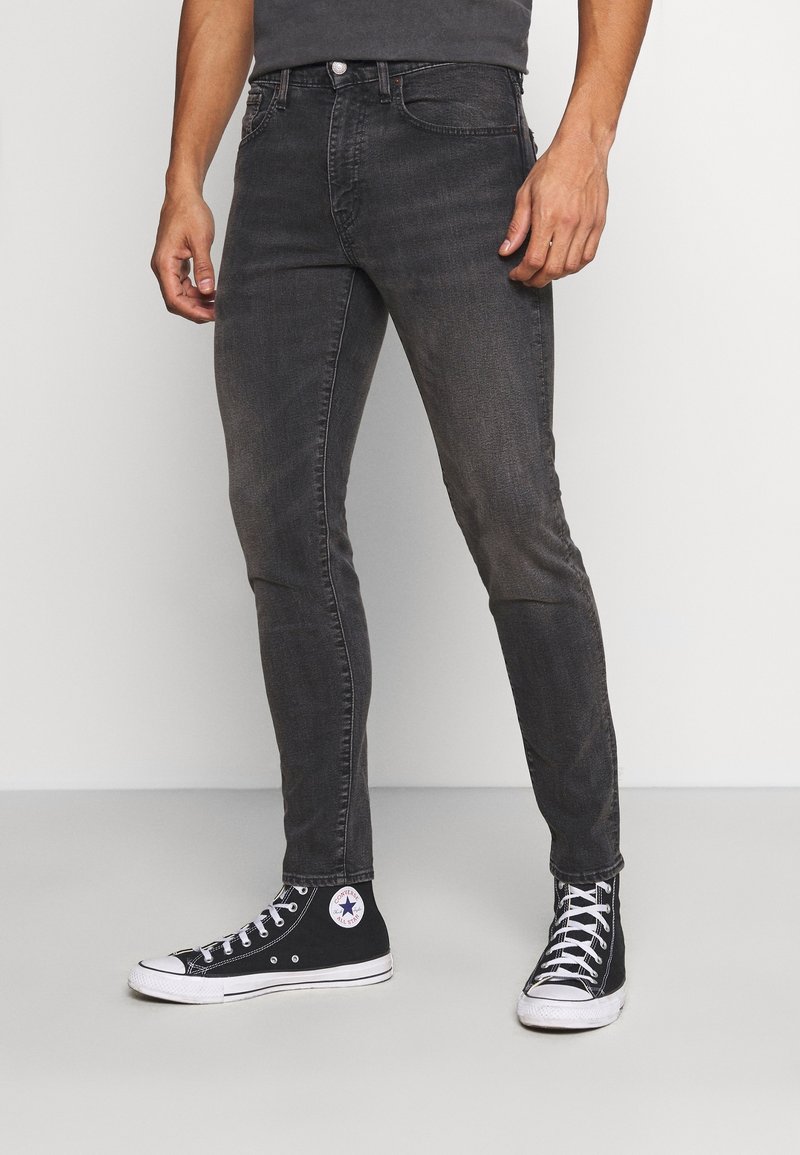 Homme portant un jean noir slim délavé et des baskets montantes noires Converse, debout sur un sol gris contre un mur blanc.