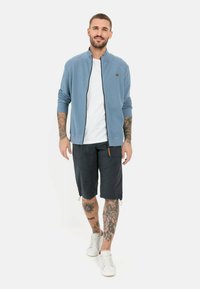 Hellblaue zip-up Jacke, weißes T-Shirt, dunkle Shorts mit Taschen und weiße Sneaker. Hat Tattoos an den Armen und Beinen.