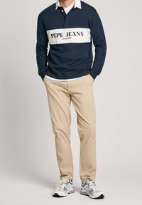 Polo a maniche lunghe blu navy con striscia bianca e testo "PEPE JEANS LONDON", abbinato a pantaloni chinos beige chiaro e sneakers grigie/bianche.