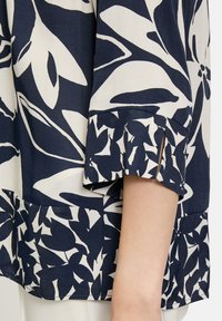 Bluse in Marineblau und Creme mit floralem Muster, kurzen Ärmeln mit Schlitzen und lockerer Passform, aus leichtem Stoff gefertigt.