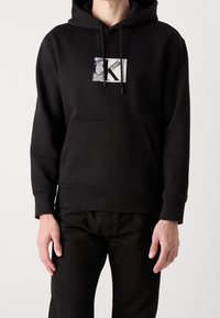 Persona vestida con una sudadera con capucha negra, que tiene un bolsillo frontal y un logo abstracto en blanco y negro "CK" en el pecho, combinada con pantalones negros.