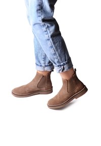 Bottines en suede marron avec des panneaux latéraux élastiques, bout arrondi et coutures contrastantes sur une semelle en caoutchouc plate. Les jeans sont légèrement retroussés aux chevilles.