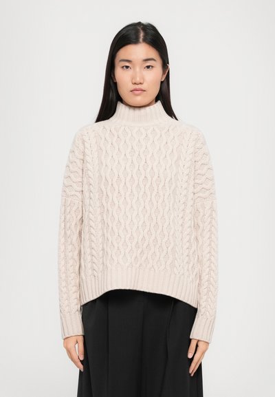 Lys beige kabelstrikket sweater med høj mock hals og ribbet kant. Har teksturerede mønstre og lange ærmer. Sammen med en sort nederdel.