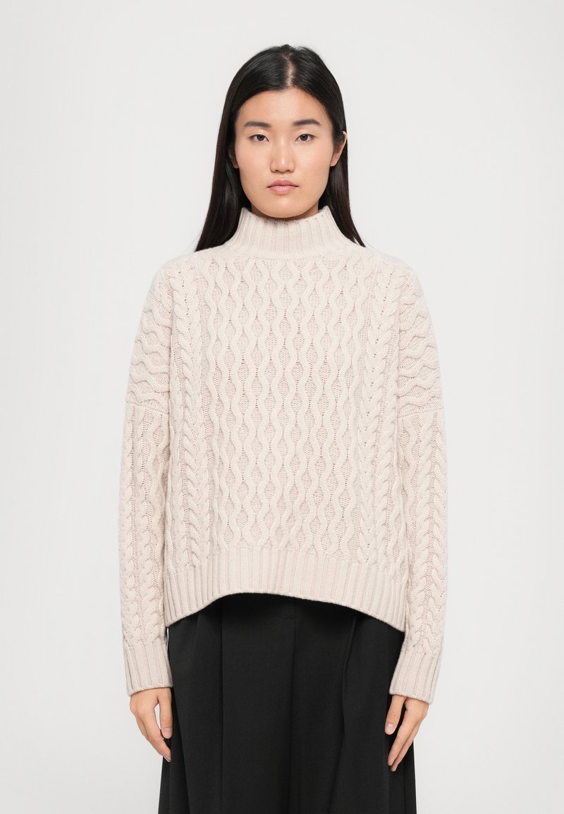Light beige cable knit sweater with a high mock neckline and ribbed hem. Features textured patterns and long sleeves. Paired with a black skirt.