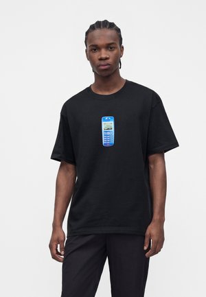 Jeune homme aux cheveux tressés portant un t-shirt noir avec un graphique bleu de téléphone mobile rétro centré sur la poitrine.